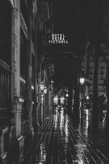 A black and white photograph of a wet, reflective street scene at night. The image features a sign for 'Hotel Reina Victoria' above a sidewalk with shiny cobblestones and large, old-fashioned street lamps casting light. Buildings line the left side, and trees are visible along the path.