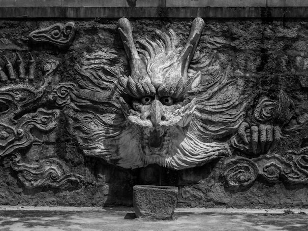 A stone carving of a dragon's face is prominently displayed on a textured wall. The dragon has large, expressive eyes with intricate details in its facial features and flowing hair, giving it a fierce and mythical appearance. Surrounding the dragon are swirling patterns that add to the mystical ambience.