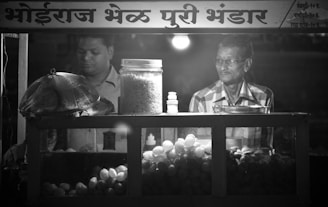 Two men are standing inside a food stall that has a variety of snacks displayed, including a large jar of puffed rice and a container of round fried snacks. One man appears to be preparing or managing the food, while the other man is looking on. The signage above the stall is written in a local script.