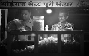 Two men are standing inside a food stall that has a variety of snacks displayed, including a large jar of puffed rice and a container of round fried snacks. One man appears to be preparing or managing the food, while the other man is looking on. The signage above the stall is written in a local script.