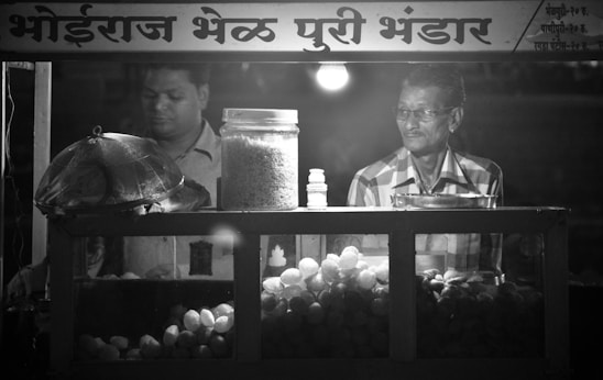 Two men are standing inside a food stall that has a variety of snacks displayed, including a large jar of puffed rice and a container of round fried snacks. One man appears to be preparing or managing the food, while the other man is looking on. The signage above the stall is written in a local script.