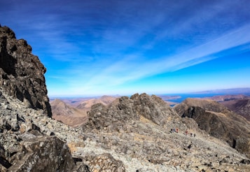 A rugged mountain landscape with rocky terrain stretches out under a clear blue sky. The distant horizon shows expansive views of surrounding peaks and a glimpse of blue water. Small groups of hikers are visible along the mountain path, emphasizing the adventurous and remote quality of the scene.
