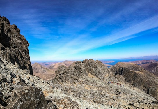 A rugged mountain landscape with rocky terrain stretches out under a clear blue sky. The distant horizon shows expansive views of surrounding peaks and a glimpse of blue water. Small groups of hikers are visible along the mountain path, emphasizing the adventurous and remote quality of the scene.
