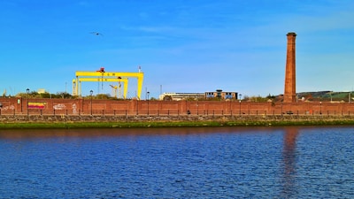 An industrial scene featuring a large yellow crane structure marked with H & W, a tall brick chimney, and various industrial buildings behind a brick wall. In the foreground, there is a calm body of water reflecting the blue sky above.