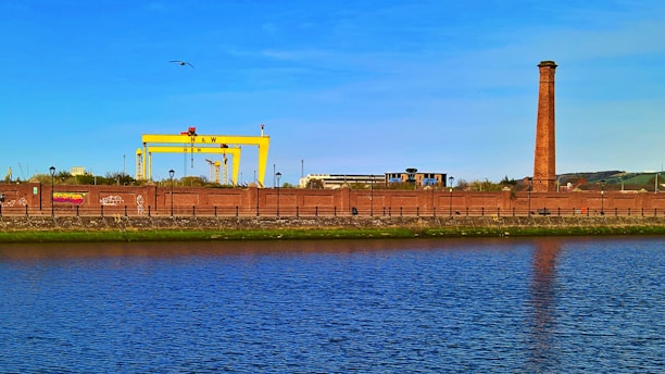 An industrial scene featuring a large yellow crane structure marked with H & W, a tall brick chimney, and various industrial buildings behind a brick wall. In the foreground, there is a calm body of water reflecting the blue sky above.