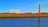 An industrial scene featuring a large yellow crane structure marked with H & W, a tall brick chimney, and various industrial buildings behind a brick wall. In the foreground, there is a calm body of water reflecting the blue sky above.