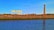 An industrial scene featuring a large yellow crane structure marked with H & W, a tall brick chimney, and various industrial buildings behind a brick wall. In the foreground, there is a calm body of water reflecting the blue sky above.
