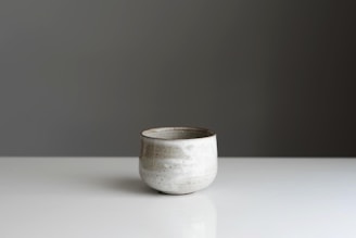 A minimalist ceramic cup is placed on a smooth, reflective surface. The cup has a simple, rounded shape with a subtle, textured pattern on its surface. The background is a plain, muted gray, enhancing the elegant simplicity of the scene.