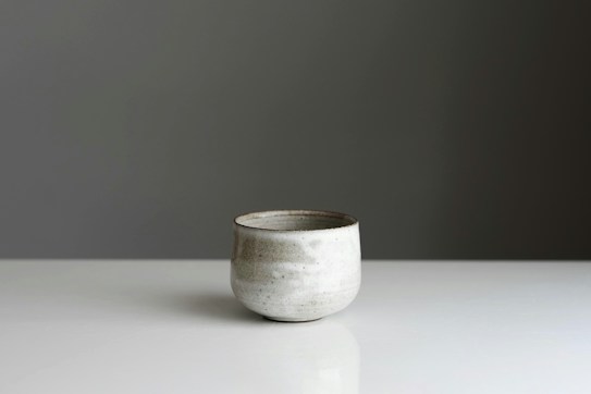 A minimalist ceramic cup is placed on a smooth, reflective surface. The cup has a simple, rounded shape with a subtle, textured pattern on its surface. The background is a plain, muted gray, enhancing the elegant simplicity of the scene.