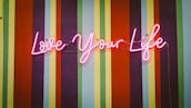Neon pink cursive lettering spells out 'Love Your Life' against a vibrant striped background, featuring alternating colors of orange, red, green, blue, and purple.