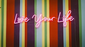Neon pink cursive lettering spells out 'Love Your Life' against a vibrant striped background, featuring alternating colors of orange, red, green, blue, and purple.