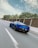 A sleek blue sports car is driving on a highway bordered by tall trees and a wall. The car speeds down the road, emphasizing motion and freedom.