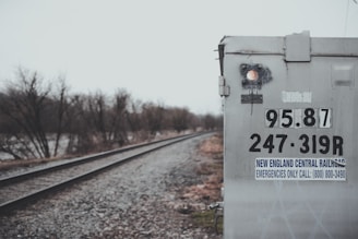 The image features a railroad track running alongside a weathered signal box or electrical cabinet. The box is labeled with numbers and a sign for the New England Central Railroad. The weather is overcast, and bare trees line the railway, suggesting a cold season.