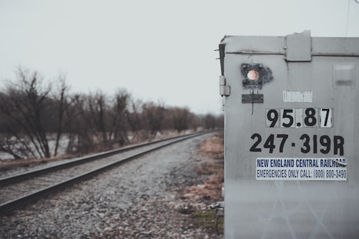 The image features a railroad track running alongside a weathered signal box or electrical cabinet. The box is labeled with numbers and a sign for the New England Central Railroad. The weather is overcast, and bare trees line the railway, suggesting a cold season.