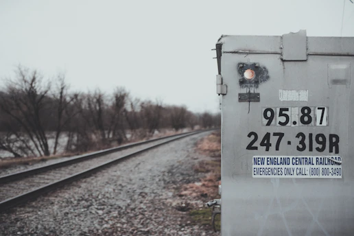 The image features a railroad track running alongside a weathered signal box or electrical cabinet. The box is labeled with numbers and a sign for the New England Central Railroad. The weather is overcast, and bare trees line the railway, suggesting a cold season.