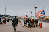 A boardwalk scene with people walking and gathering around a vendor stall, which is adorned with Puerto Rican flags. The environment suggests a public space, possibly in an urban area, with stadium lights and city buildings visible in the background. A mix of people are visible, some standing in groups, while others are walking away or towards the camera.