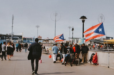 A boardwalk scene with people walking and gathering around a vendor stall, which is adorned with Puerto Rican flags. The environment suggests a public space, possibly in an urban area, with stadium lights and city buildings visible in the background. A mix of people are visible, some standing in groups, while others are walking away or towards the camera.