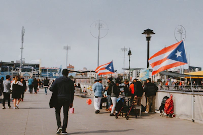 A boardwalk scene with people walking and gathering around a vendor stall, which is adorned with Puerto Rican flags. The environment suggests a public space, possibly in an urban area, with stadium lights and city buildings visible in the background. A mix of people are visible, some standing in groups, while others are walking away or towards the camera.