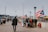 A boardwalk scene with people walking and gathering around a vendor stall, which is adorned with Puerto Rican flags. The environment suggests a public space, possibly in an urban area, with stadium lights and city buildings visible in the background. A mix of people are visible, some standing in groups, while others are walking away or towards the camera.