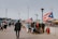 A boardwalk scene with people walking and gathering around a vendor stall, which is adorned with Puerto Rican flags. The environment suggests a public space, possibly in an urban area, with stadium lights and city buildings visible in the background. A mix of people are visible, some standing in groups, while others are walking away or towards the camera.
