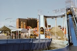 An amusement park ride featuring a water slide is seen in a lively setting. The ride, labeled 'Wild River,' includes a steep incline where people are descending on log boats. The background consists of various amusement park structures, colorful tents, and a multi-story building.