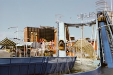 An amusement park ride featuring a water slide is seen in a lively setting. The ride, labeled 'Wild River,' includes a steep incline where people are descending on log boats. The background consists of various amusement park structures, colorful tents, and a multi-story building.