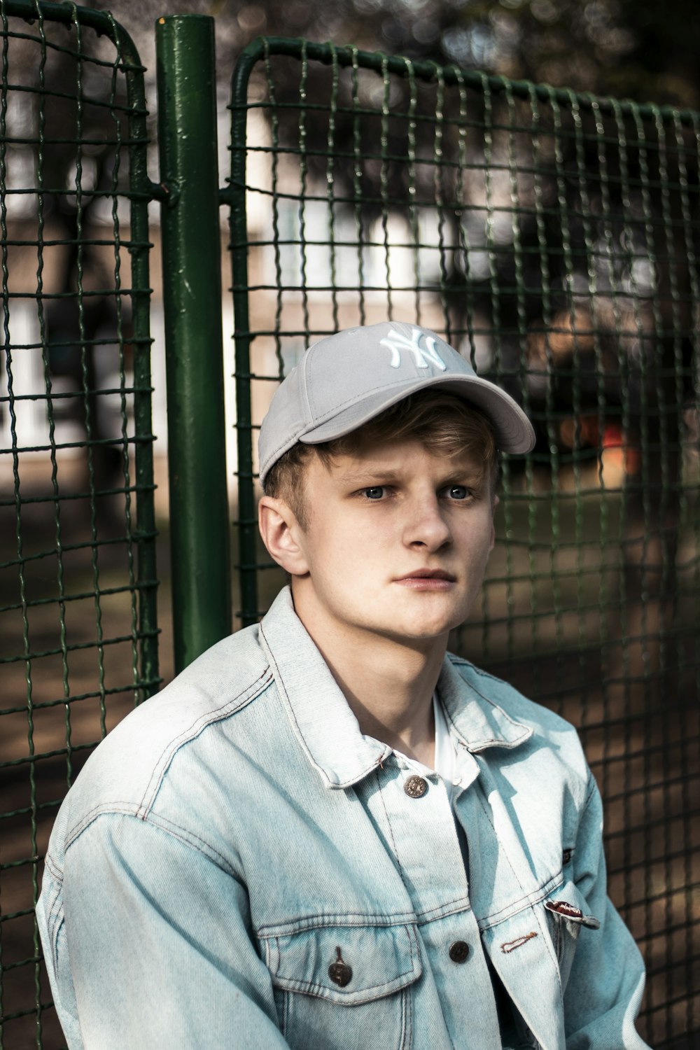 man wearing blue button-up denim jacket sitting beside green fence