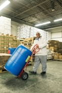 A person in a warehouse is transporting a large blue barrel on a red hand truck. The background includes stacks of cardboard boxes and pallets, with fluorescent lighting from above and a concrete floor.