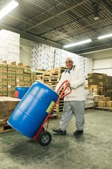 A person in a warehouse is transporting a large blue barrel on a red hand truck. The background includes stacks of cardboard boxes and pallets, with fluorescent lighting from above and a concrete floor.