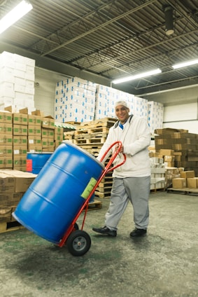 A person in a warehouse is transporting a large blue barrel on a red hand truck. The background includes stacks of cardboard boxes and pallets, with fluorescent lighting from above and a concrete floor.