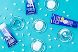 A flat lay composition featuring two blue tubes of skincare products and transparent dishes containing cream on a bright blue background. White oval pills are scattered throughout the scene, adding texture and contrast.