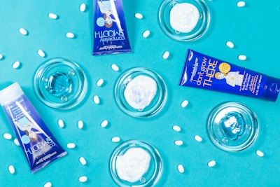 A flat lay composition featuring two blue tubes of skincare products and transparent dishes containing cream on a bright blue background. White oval pills are scattered throughout the scene, adding texture and contrast.