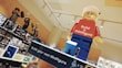 A large LEGO minifigure model with a red shirt that says 'Build a minifigure' and blue pants is displayed prominently in a retail environment. Shelves in the background are filled with various LEGO sets and boxes. Some of the LEGO sets feature characters from different themes and franchises.
