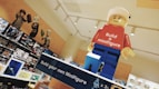A large LEGO minifigure model with a red shirt that says 'Build a minifigure' and blue pants is displayed prominently in a retail environment. Shelves in the background are filled with various LEGO sets and boxes. Some of the LEGO sets feature characters from different themes and franchises.