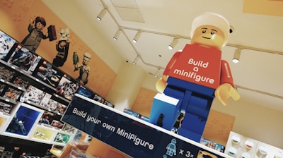 A large LEGO minifigure model with a red shirt that says 'Build a minifigure' and blue pants is displayed prominently in a retail environment. Shelves in the background are filled with various LEGO sets and boxes. Some of the LEGO sets feature characters from different themes and franchises.