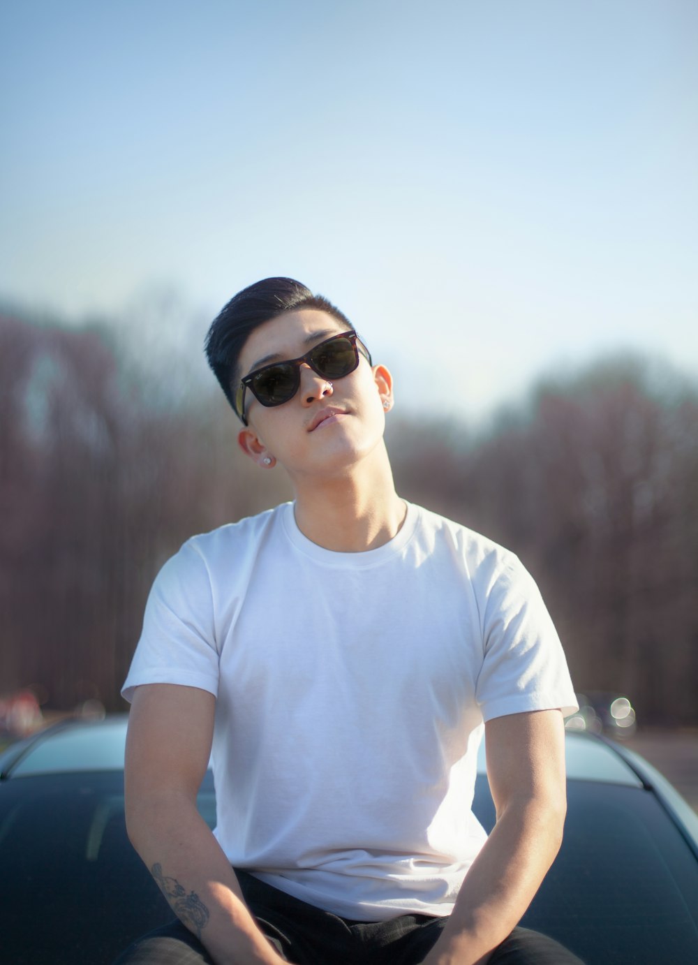 man wearing plain white shirt and black wayfarer sunglasses sitting on hood of car