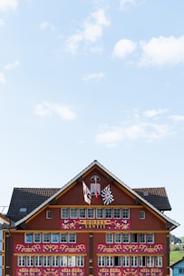 A traditional Swiss-style hotel facade adorned with intricate decorative elements, featuring two flags and multiple windows. The building is painted in earthy red tones with white details. The roof is lined with dark shingles, and the sky is bright with scattered clouds.