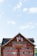 A traditional Swiss-style hotel facade adorned with intricate decorative elements, featuring two flags and multiple windows. The building is painted in earthy red tones with white details. The roof is lined with dark shingles, and the sky is bright with scattered clouds.