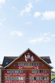 A traditional Swiss-style hotel facade adorned with intricate decorative elements, featuring two flags and multiple windows. The building is painted in earthy red tones with white details. The roof is lined with dark shingles, and the sky is bright with scattered clouds.