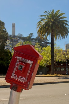 A red fire alarm box is prominently displayed in the foreground, mounted on a metal pole. In the background, there is a tall, white tower situated on a hill surrounded by trees and residential buildings. A large palm tree is visible to the right of the fire alarm box, adding to the urban landscape.