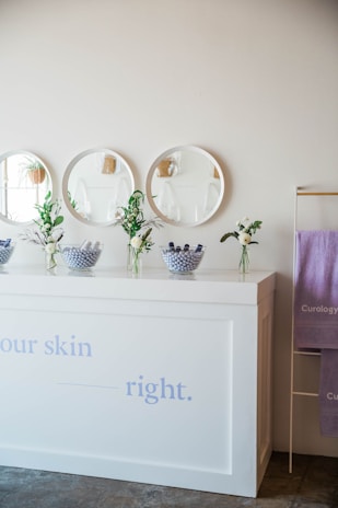 A minimalist interior setup featuring a white counter adorned with small vases of fresh flowers and bowls filled with purple bath bombs. Above the counter, there are three round mirrors mounted on a clean, neutral wall. To the side, a metal rack holds purple towels with branding.