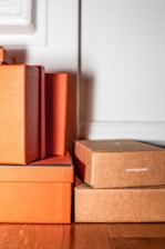 A stack of rectangular boxes in orange and brown shades is positioned against a light-colored wall. The texture of the boxes is slightly textured, suggesting they are made of cardboard or a similar material. Some boxes are marked with the text 'curology.com'. They are placed on a wooden floor, adding a warm tone to the scene.