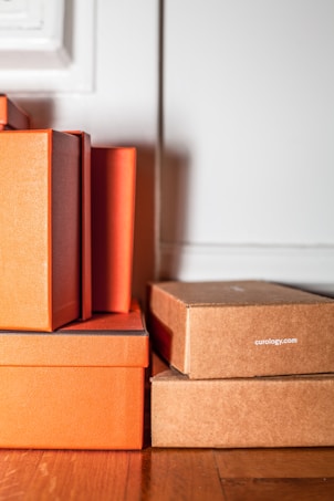 A stack of rectangular boxes in orange and brown shades is positioned against a light-colored wall. The texture of the boxes is slightly textured, suggesting they are made of cardboard or a similar material. Some boxes are marked with the text 'curology.com'. They are placed on a wooden floor, adding a warm tone to the scene.
