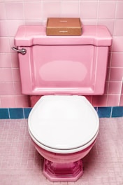 A pink toilet with a white seat is set against pink tiled walls and floor. On top of the toilet tank, there is a small, brown cardboard box.