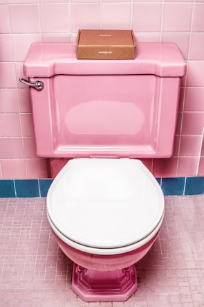 A pink toilet with a white seat is set against pink tiled walls and floor. On top of the toilet tank, there is a small, brown cardboard box.