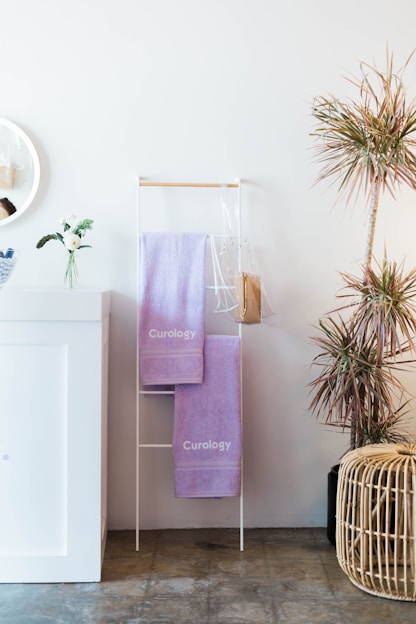 A minimalist bathroom setup includes a white cabinet with a small vase holding white flowers and greenery. Beside it, a slim rack holds two purple towels labeled 'Curology'. A circular mirror partly visible adds elegance to the decor. To the right, a potted plant with thin, spiky leaves provides a touch of nature, and a woven basket sits on the floor nearby.