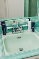 A vintage-style bathroom sink with light green tiles and chrome fixtures. Several skincare product bottles are placed on the counter around the sink.