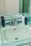 A vintage-style bathroom sink with light green tiles and chrome fixtures. Several skincare product bottles are placed on the counter around the sink.