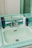 A vintage-style bathroom sink with light green tiles and chrome fixtures. Several skincare product bottles are placed on the counter around the sink.