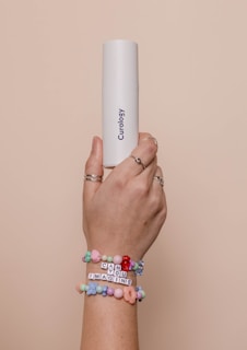 A hand with multiple rings on the fingers is holding a white cylindrical container labeled 'Curology'. The wrist is adorned with colorful beaded bracelets, one of which spells out 'CAN YOU IMAGINE'. The background is a soft beige color, creating a neutral setting.
